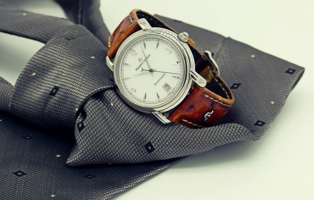 Une montre Maurice Lacroix à cadran blanc et bracelet en cuir marron posée sur une cravate grise à motifs.